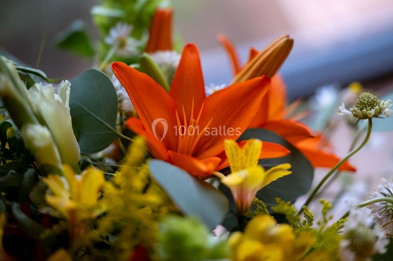 Fleur de lys orange entourée de feuillage vert et de petites fleurs jaunes et blanches dans un bouquet lumineux.