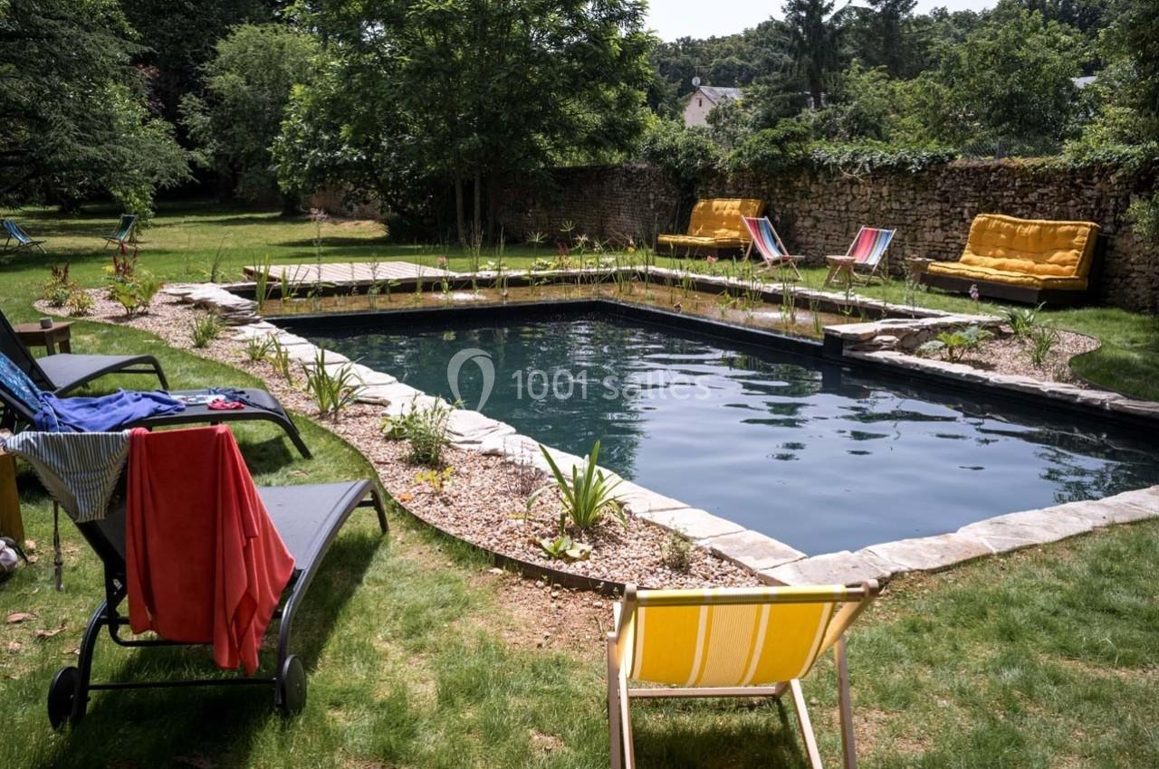Piscine naturelle entourée de chaises longues, végétation et murs en pierre dans un jardin verdoyant.
