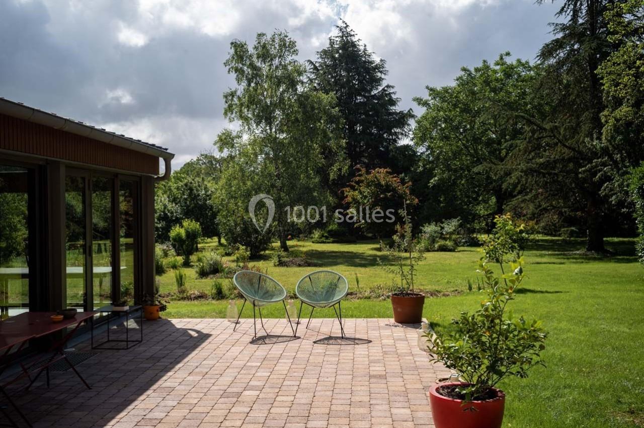 Terrasse en pavés avec deux fauteuils, entourée de plantes en pots, donnant sur un grand jardin arboré.