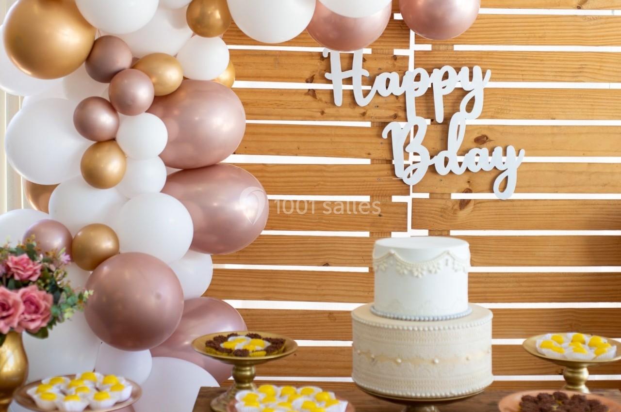 Décoration d'anniversaire avec ballons blancs et dorés, gâteau central blanc et desserts sur une table en bois.