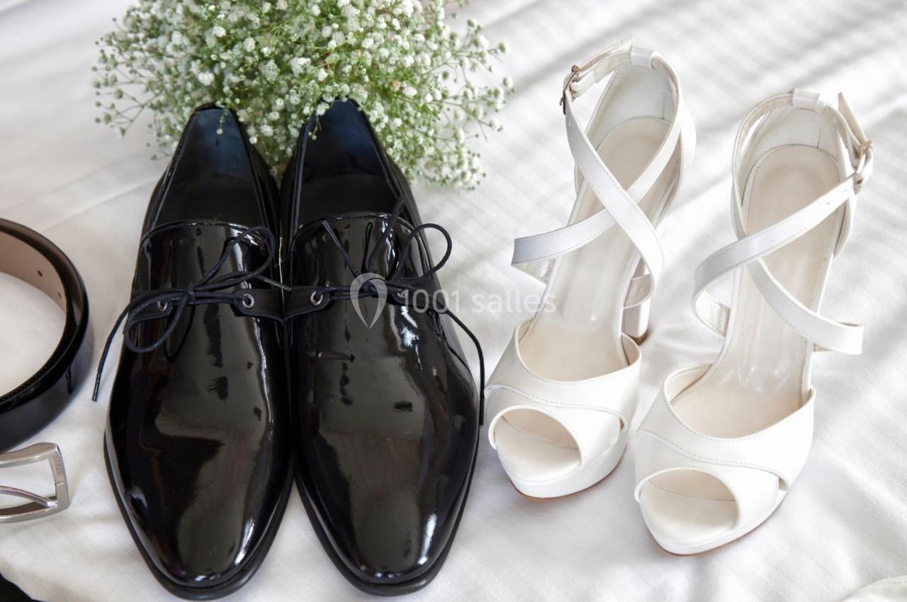 Chaussures noires vernies pour homme et sandales blanches à talons pour femme posées sur un lit avec un bouquet de fleurs.