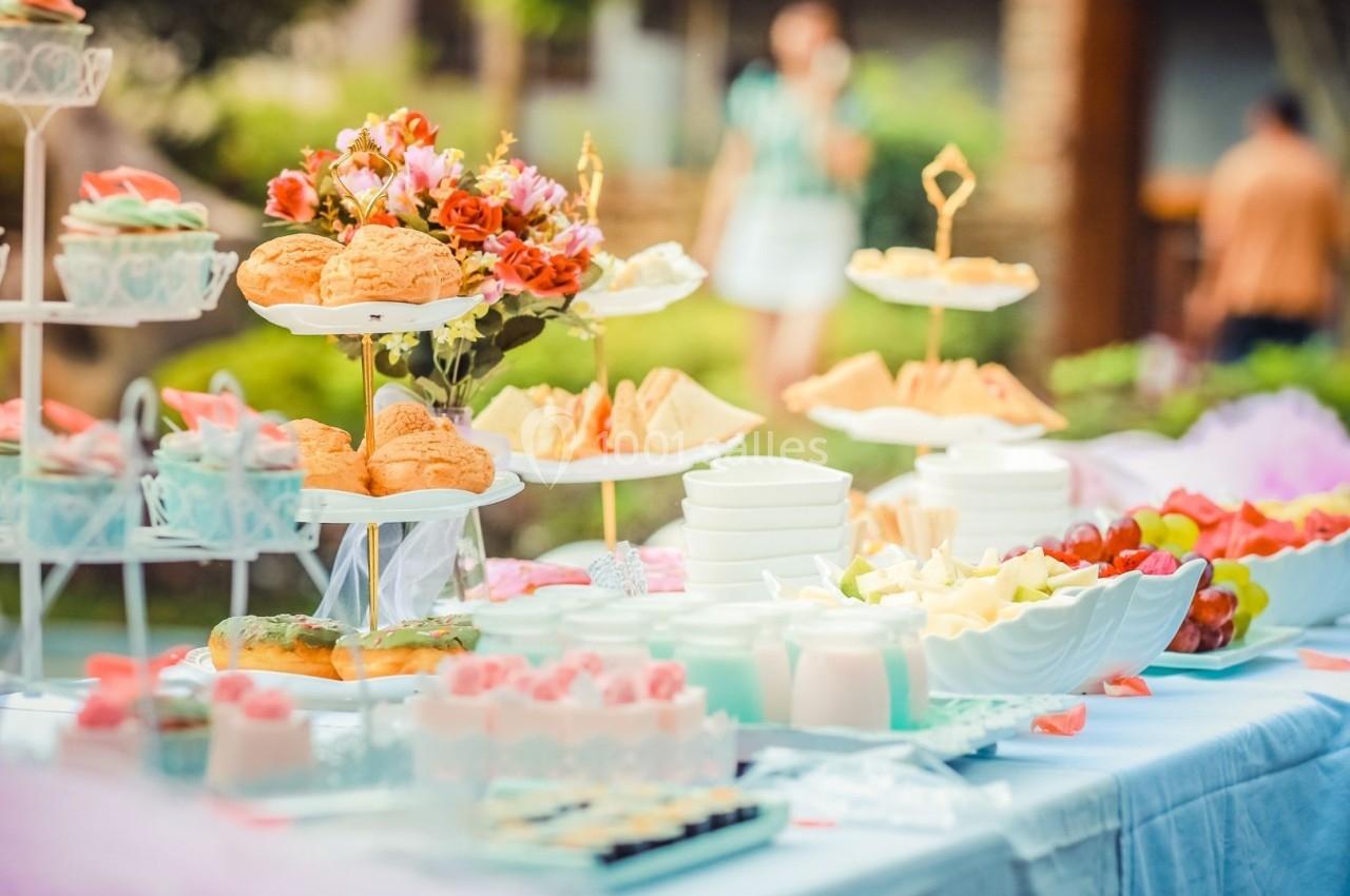 Table dressée en extérieur avec des desserts, fruits, sandwichs et boissons, décorée de fleurs et de couleurs pastel.