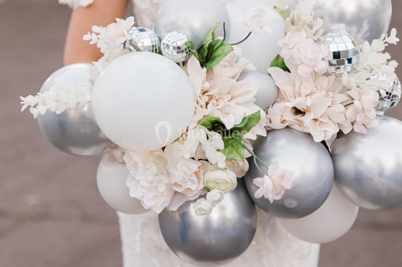 Bouquet composé de fleurs blanches, de ballons argentés et blancs, et de petites décorations en forme de boules miroir.