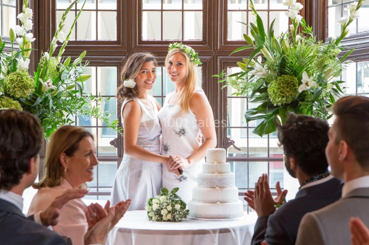 Deux femmes en robe blanche sourient devant un gâteau de mariage, entourées d'invités applaudissant dans une salle lumineuse.