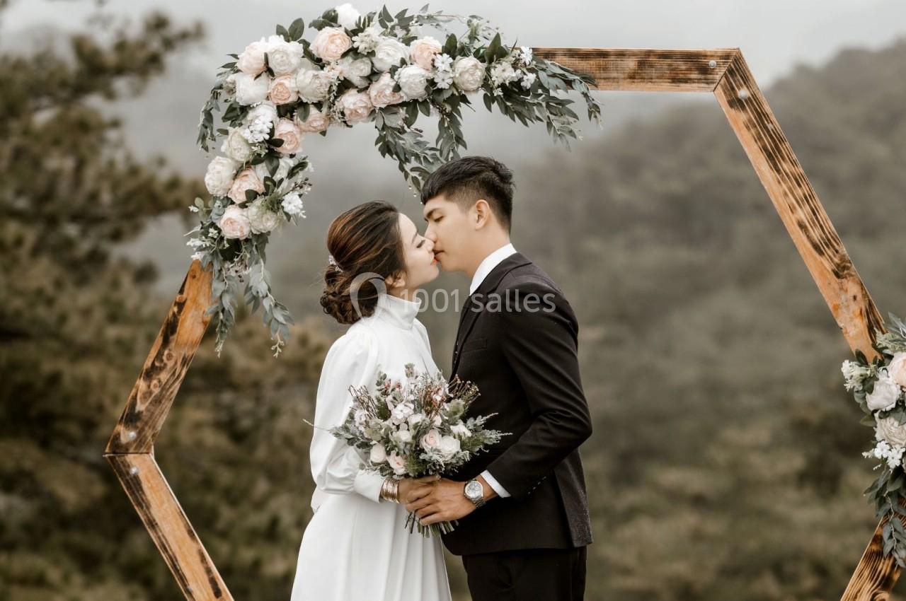 Un couple échange un baiser sous une arche hexagonale décorée de fleurs, avec un paysage naturel en arrière-plan.
