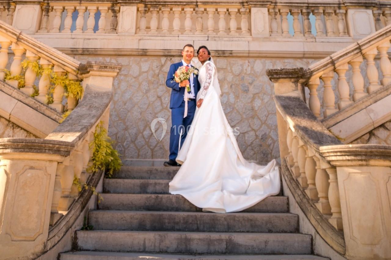 Un couple de mariés pose sur un escalier en pierre devant une balustrade ornée, dans un cadre élégant et ensoleillé.
