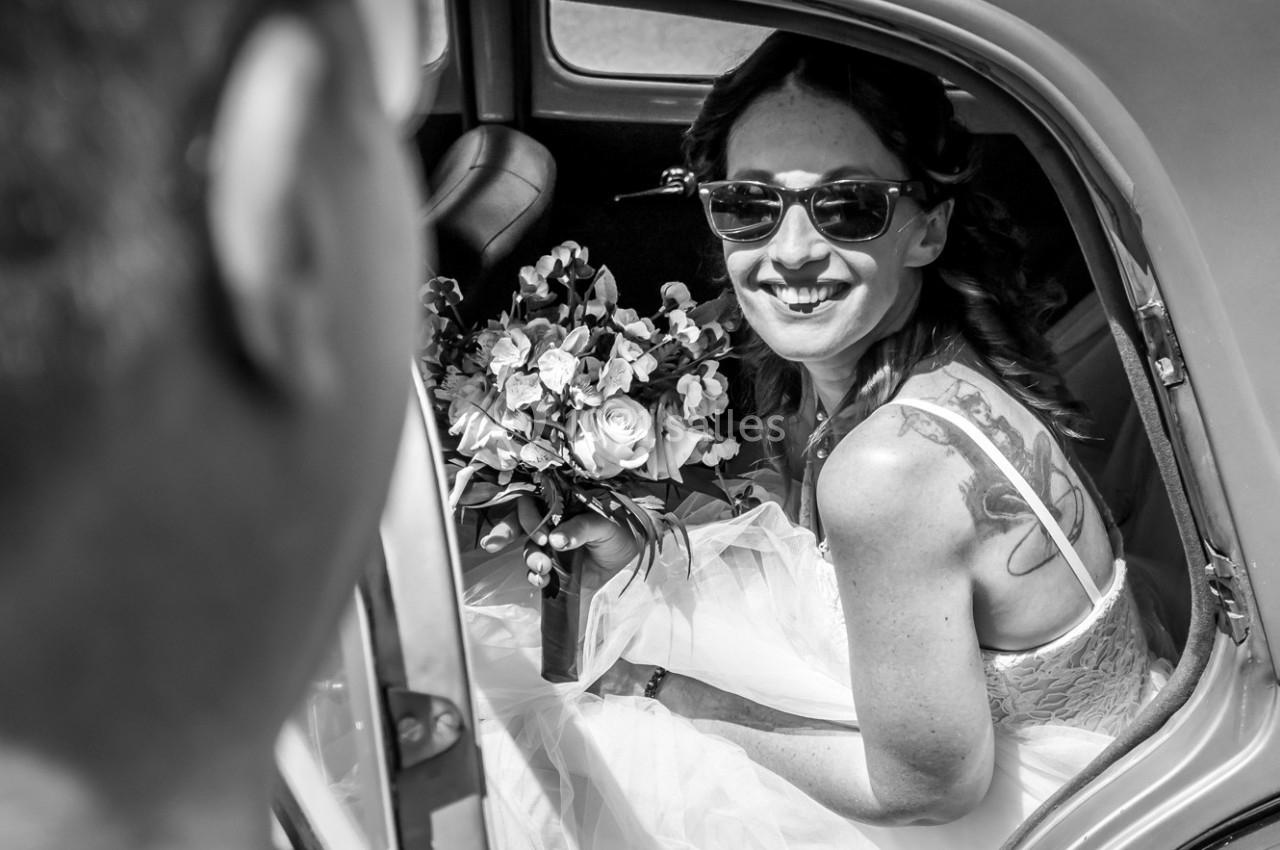 Une mariée souriante, portant des lunettes de soleil, tient un bouquet de fleurs dans une voiture ancienne.