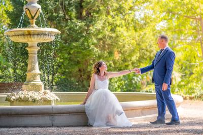 Femme en robe blanche souriante, assise près d'une fontaine, avec un homme en costume visible de dos.