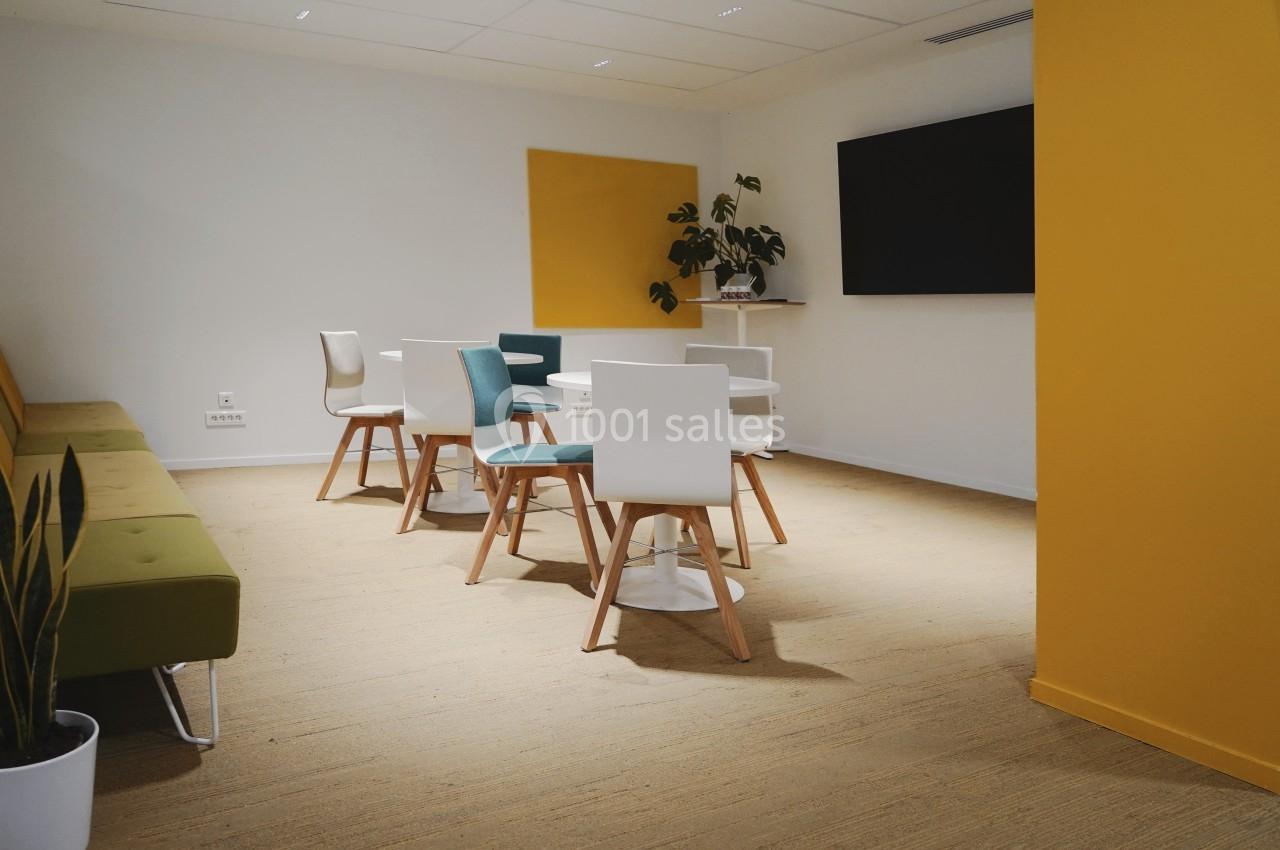 Salle lumineuse avec des chaises colorées autour de petites tables, un canapé vert et un écran mural.