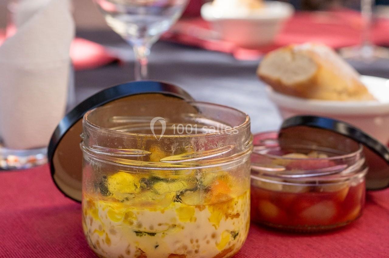 Deux bocaux en verre contenant des plats, posés sur une table dressée avec une nappe rouge et des verres.