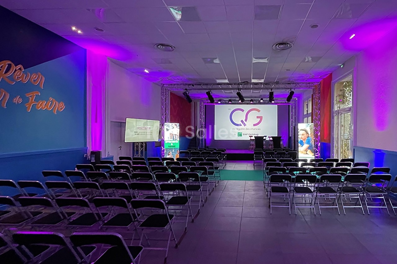 Salle de conférence avec rangées de chaises vides, écran de projection allumé et éclairage violet.