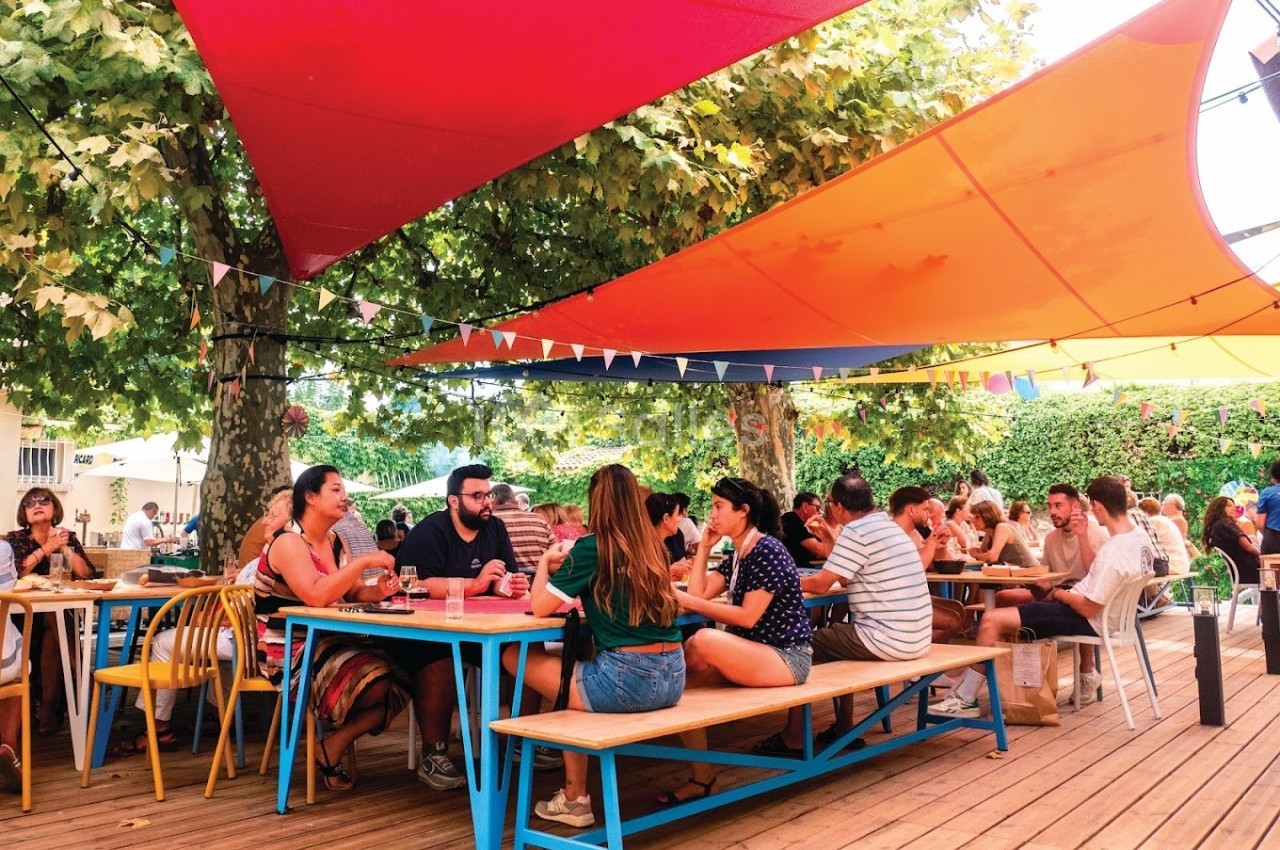 Des personnes discutent et partagent un repas à des tables en bois sous des voiles d'ombrage colorées en extérieur.