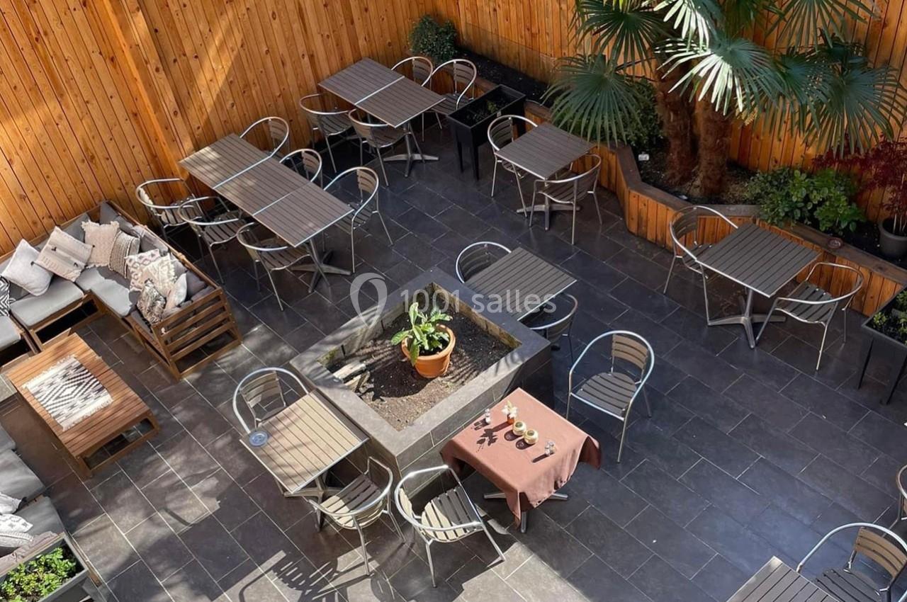 Terrasse extérieure avec tables, chaises en métal, plantes et coin salon sur un sol en dalles sombres.