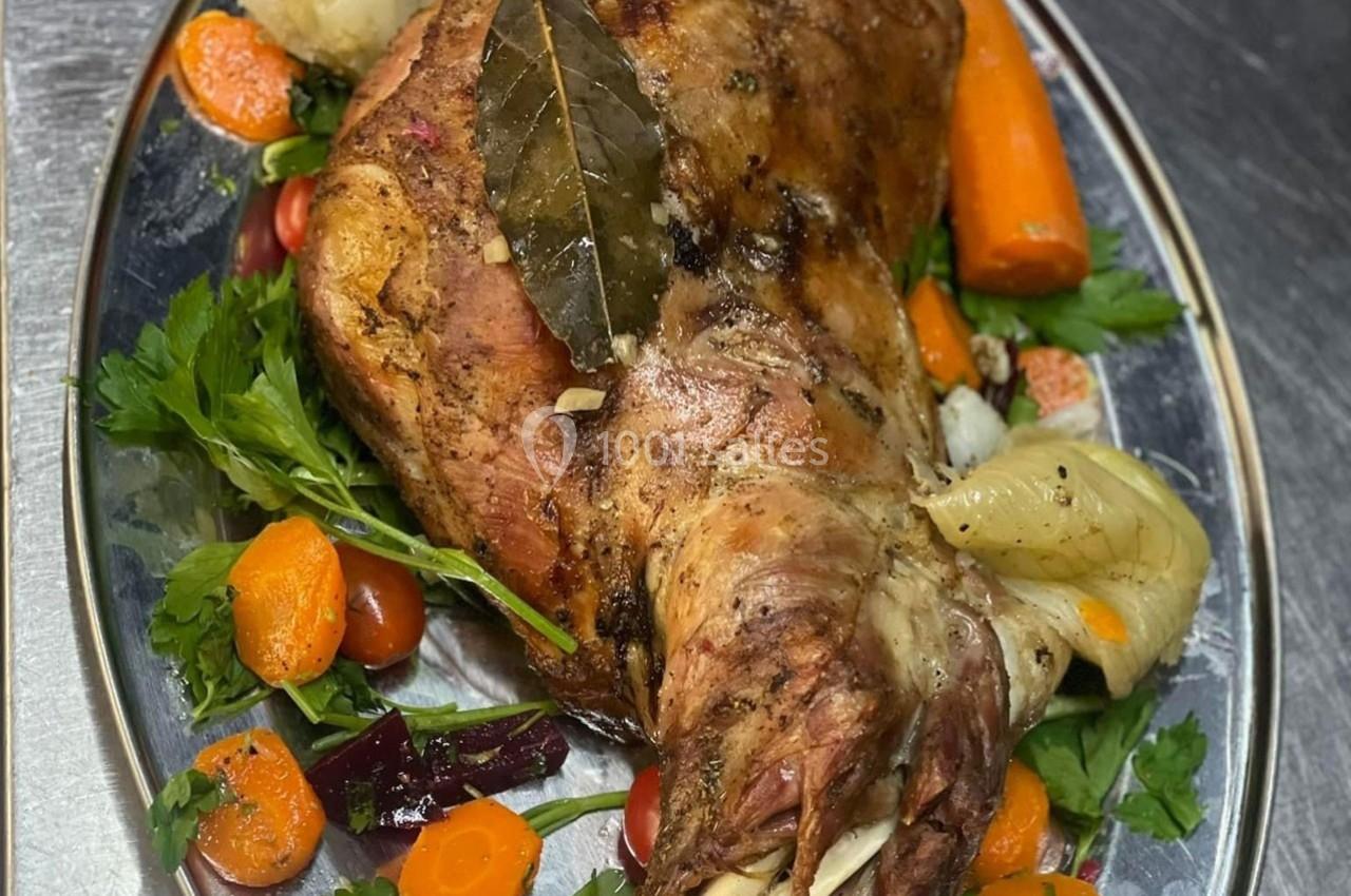 Gigot d'agneau rôti garni de légumes colorés, présenté sur un plat en métal.