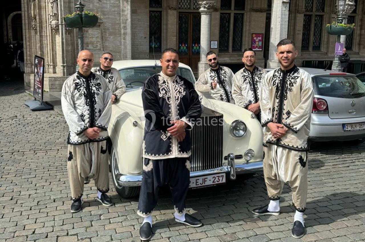 Six hommes en tenue traditionnelle posent devant une voiture ancienne blanche sur une place pavée.