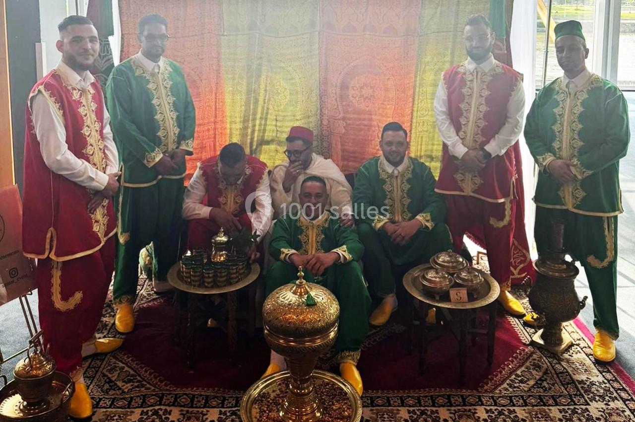 Des hommes en tenues traditionnelles colorées posent autour d'objets décoratifs sur un tapis dans un décor oriental.