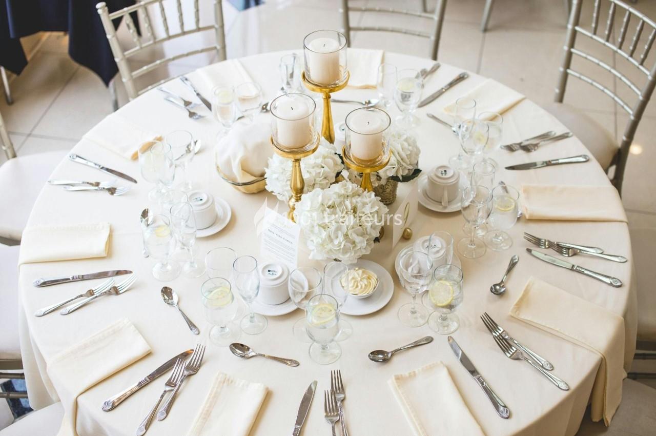 Table ronde élégamment dressée avec nappes blanches, couverts, verres, bougies et centre de table floral.