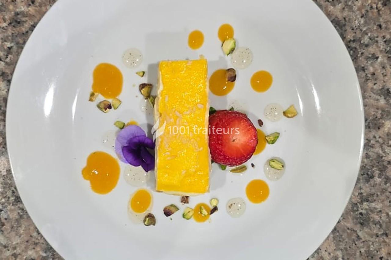 Dessert rectangulaire jaune avec fraise, pistaches, fleurs comestibles et sauces colorées sur une assiette blanche.