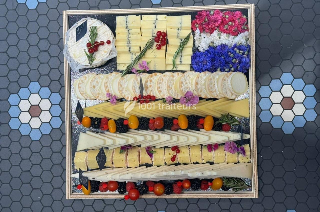 Plateau de fromages variés joliment disposés avec fleurs comestibles, fruits rouges et tomates cerises sur fond de carrelage.