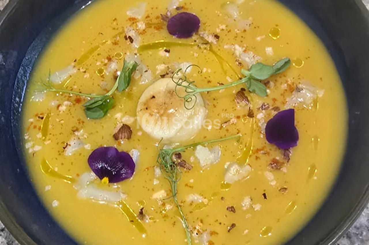 Soupe de légumes jaune garnie de fleurs comestibles, herbes fraîches, noix concassées et filet d'huile.