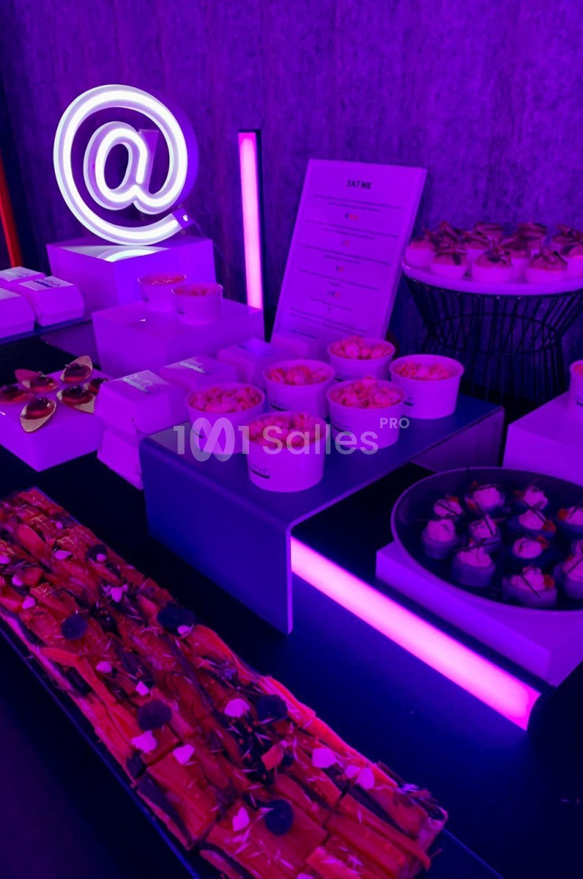 Comptoir de dégustation aux néons rose et violet - enseigne arrobe lumineuse, coupes de cocktails et petits-fours.