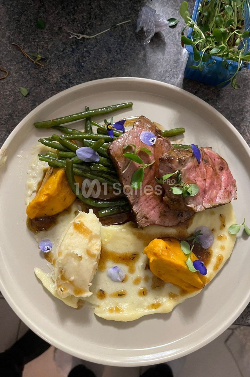 Assiette avec viande rosée, haricots verts, purée et légumes jaunes décorés de fleurs comestibles.