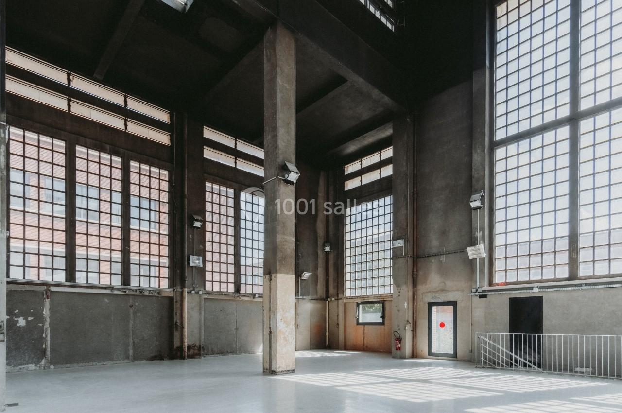 Intérieur d'un grand bâtiment industriel avec de hauts plafonds, des colonnes en béton et de larges fenêtres quadrillées.