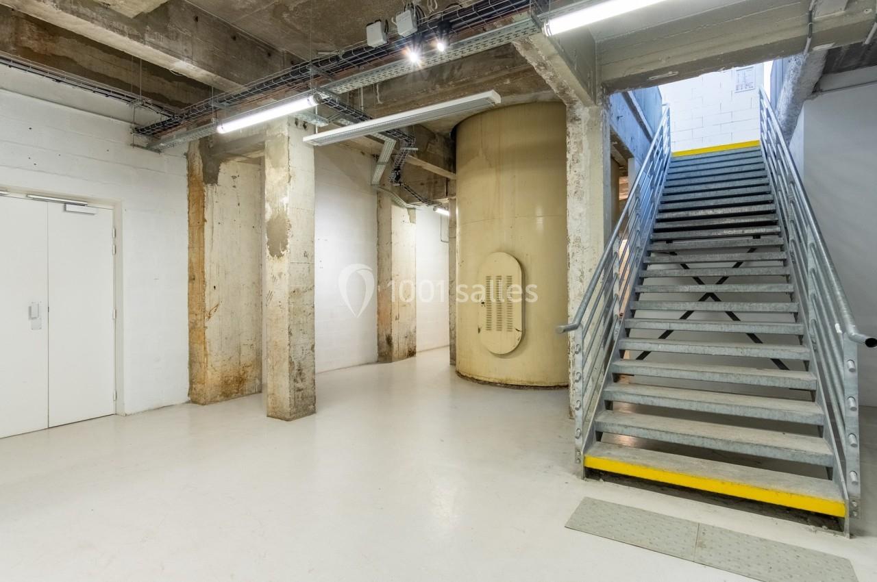 Escalier métallique montant à droite, murs en béton brut et réservoir cylindrique beige dans un espace industriel.