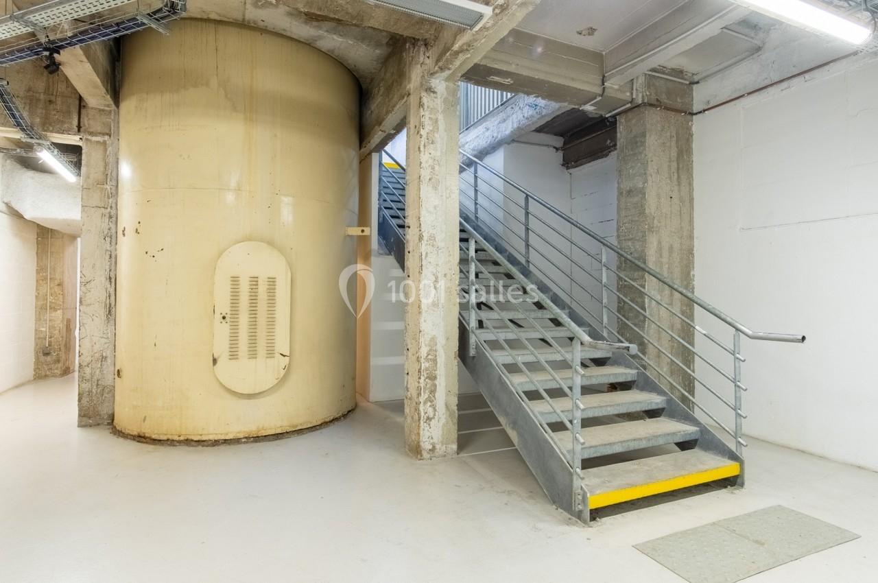 Escalier métallique avec rampe, situé dans un espace industriel avec un grand réservoir cylindrique beige.