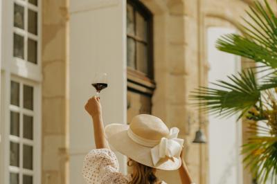 Miniature Location salle Margaux (Gironde) - Château Pontac Lynch #7 Femme de dos, vêtue d'un chapeau et d'une tenue claire, levant un verre de vin devant une façade en pierre avec volets…