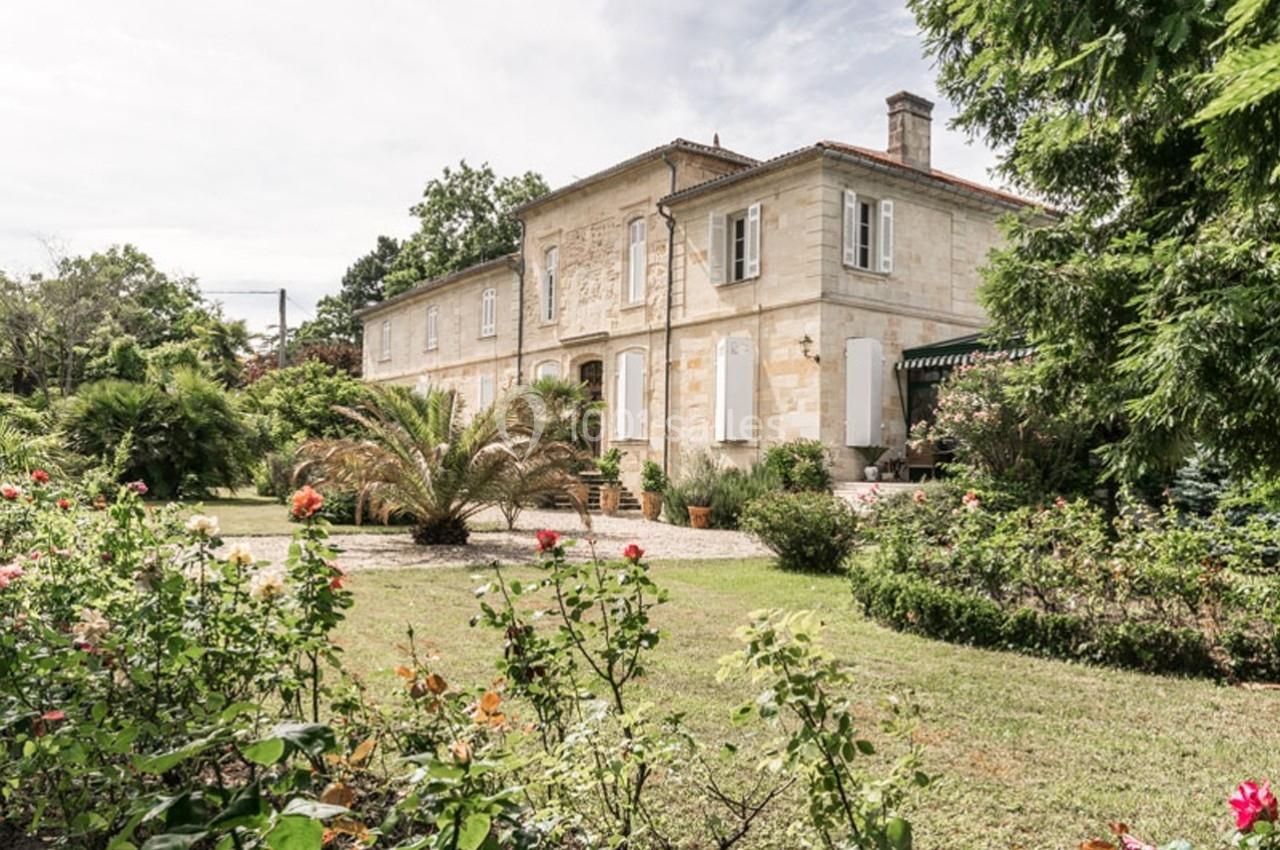 Maison en pierre entourée d'un jardin fleuri avec des palmiers et des rosiers, sous un ciel légèrement nuageux.
