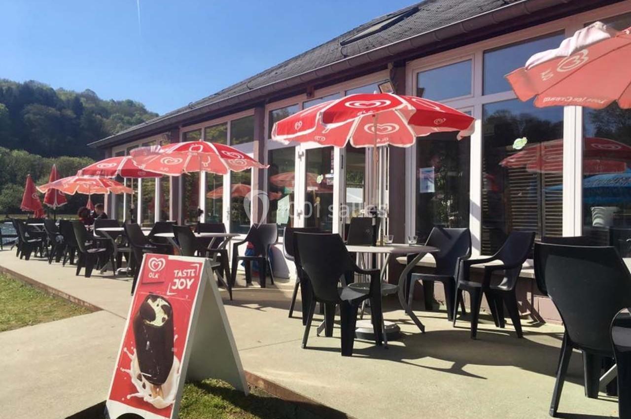 Terrasse ensoleillée avec des tables, chaises noires et parasols rouges, devant un bâtiment vitré.