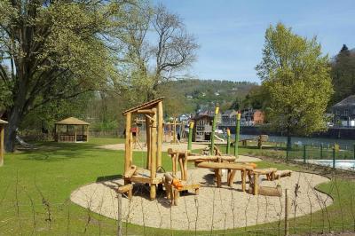 Aire de jeux en bois sur un sol sablé, entourée de verdure et située près d'une rivière et de collines.