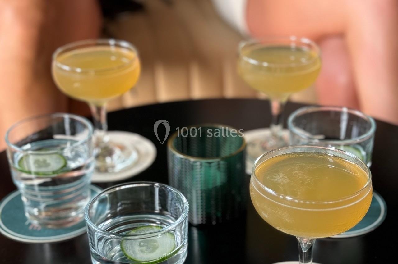 Verres de cocktails jaunes et d'eau avec rondelles de concombre posés sur une table noire, entourés de convives.