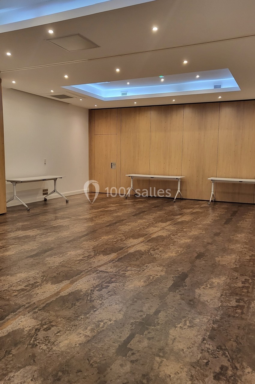 Salle vide avec parquet sombre, murs en bois clair, plafond lumineux et deux tables pliantes contre le mur.