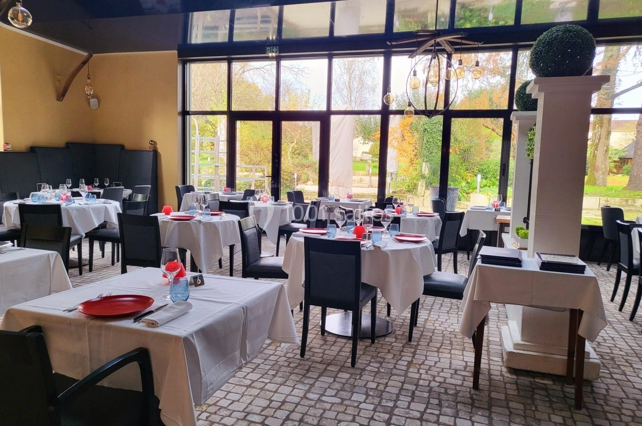 Salle de restaurant lumineuse avec grandes baies vitrées, tables dressées avec nappes blanches et chaises noires.