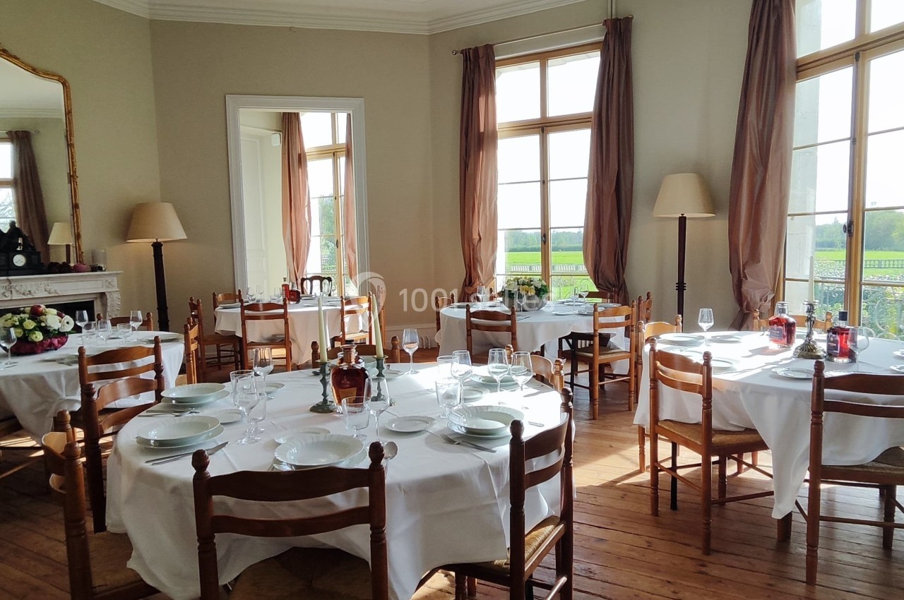 Salle lumineuse avec tables dressées, nappes blanches, chaises en bois, grandes fenêtres et vue sur un paysage verdoyant.