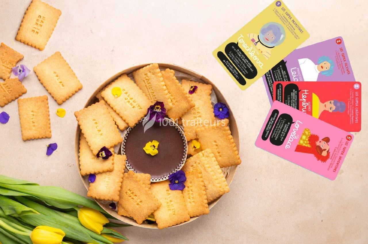 Assortiment de biscuits autour d'une ganache au chocolat, décorés de fleurs comestibles, avec des cartes colorées à côté.
