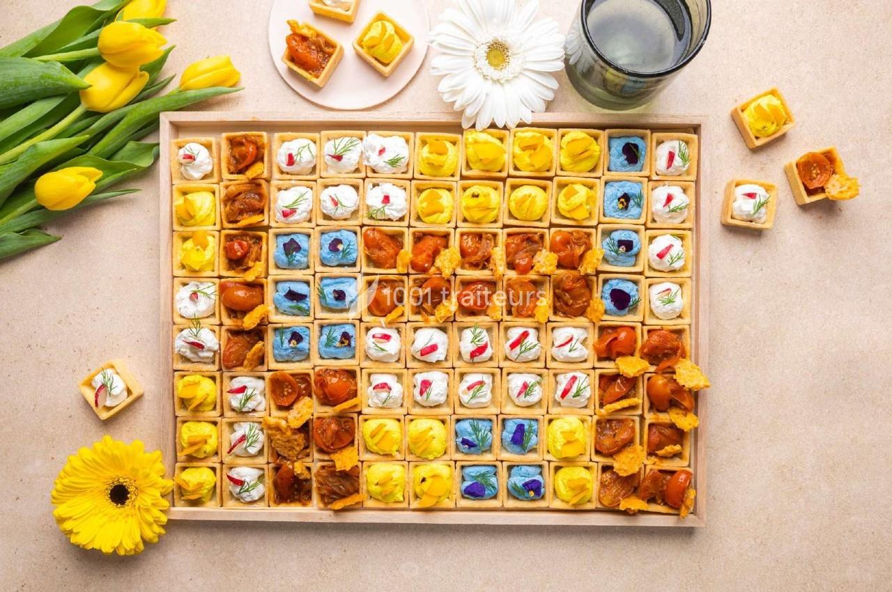 Plateau de bouchées apéritives colorées disposées en rangées, entouré de fleurs jaunes et d'un verre d'eau.