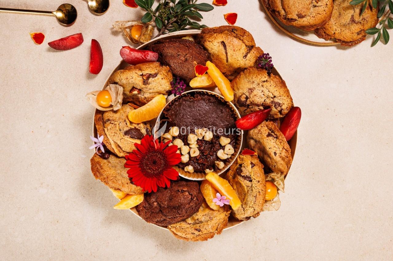 Assortiment de cookies et de fruits disposés autour d'un gâteau au chocolat, décoré de fleurs et de noix.
