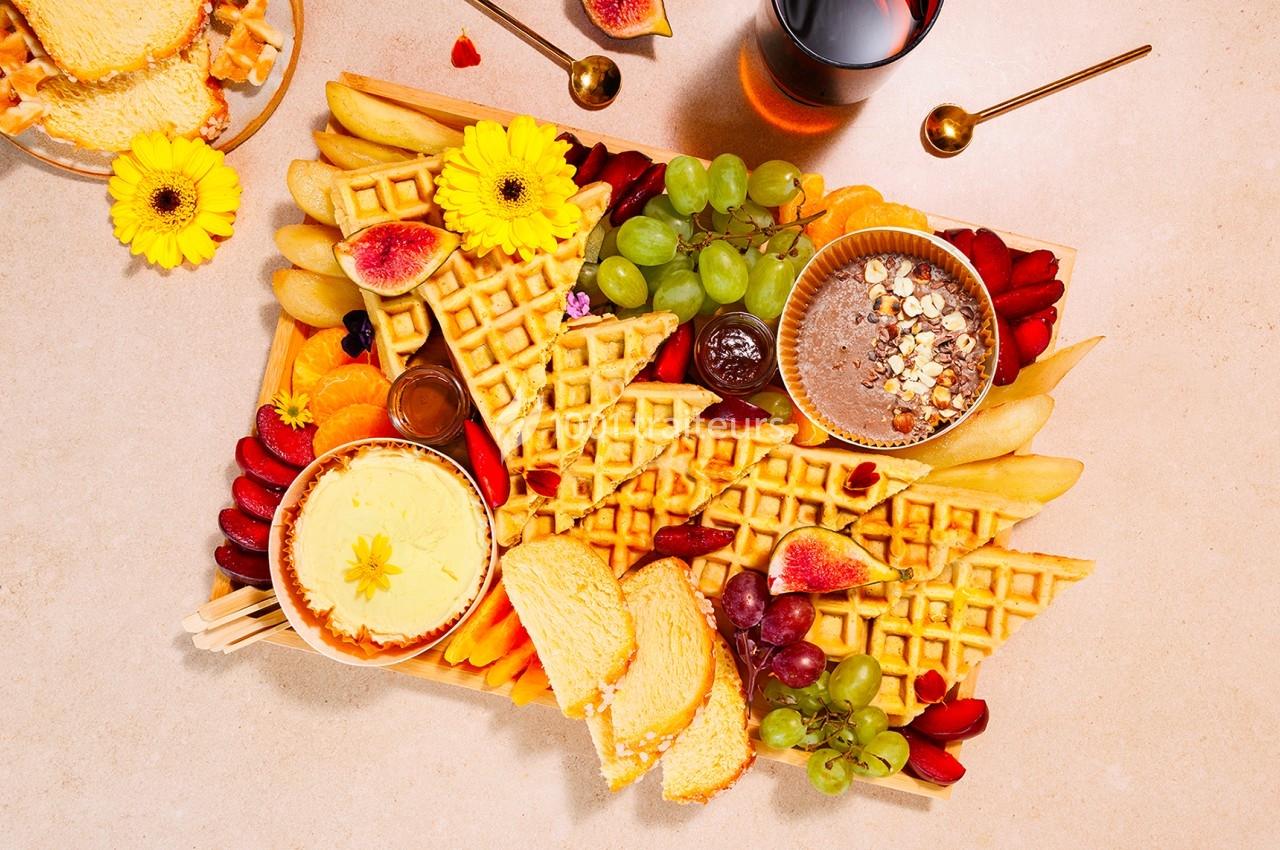 Plateau garni de gaufres, fruits frais, tartinades, fleurs décoratives et boissons sur une table beige.