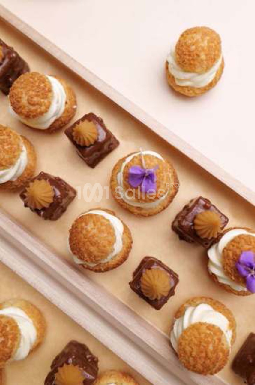 Petits fours sur plateau en bois : choux garnis de crème blanche, cubes au chocolat ornés d'une rosette dorée et fleurs violettes comestibles. Let me check length: roughly 150 characters, too long. Need ≤125. Petits fours sur plateau en bois : choux à la