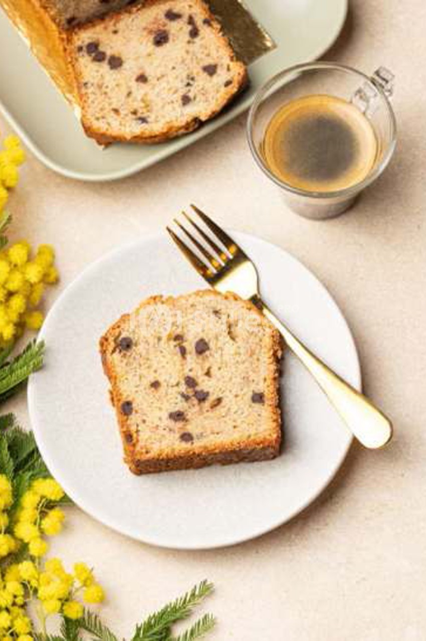 Tranche de cake aux pépites de chocolat posée sur une assiette blanche, à côté d'une fourchette dorée, d'un verre de café et d'un cake entier en arrière-plan.