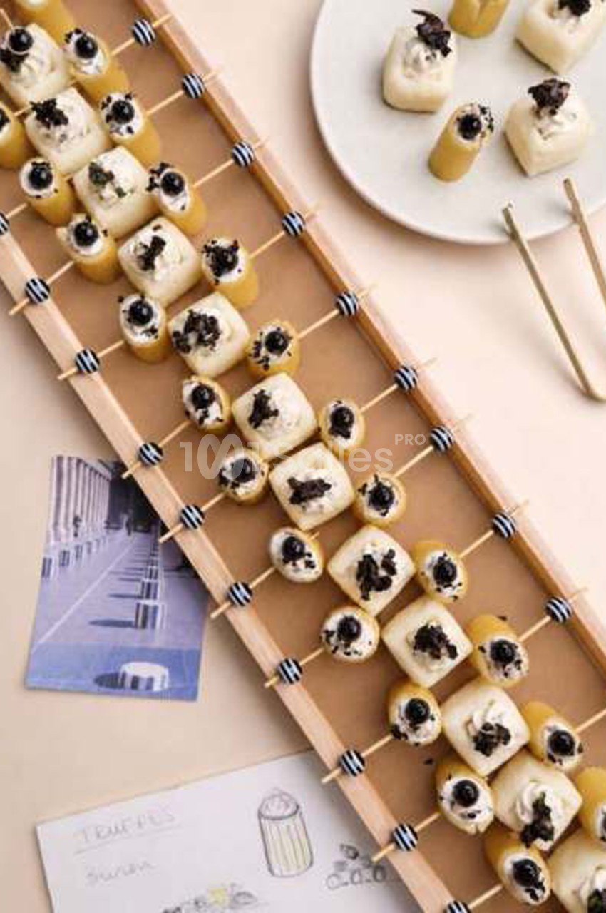 Bouchées apéritives sur piques en bambou disposées en diagonale sur une planche en bois, cubes blancs garnis de miettes sombres.