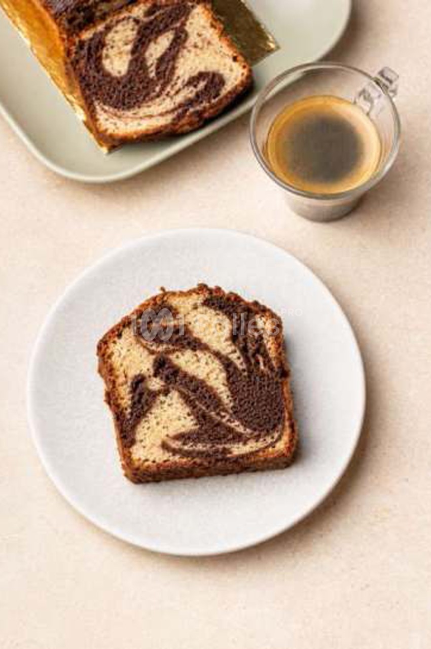 Tranche de cake marbré chocolat et vanille posée sur une assiette blanche, à côté d'un verre de café et d'un plat contenant le reste du gâteau.