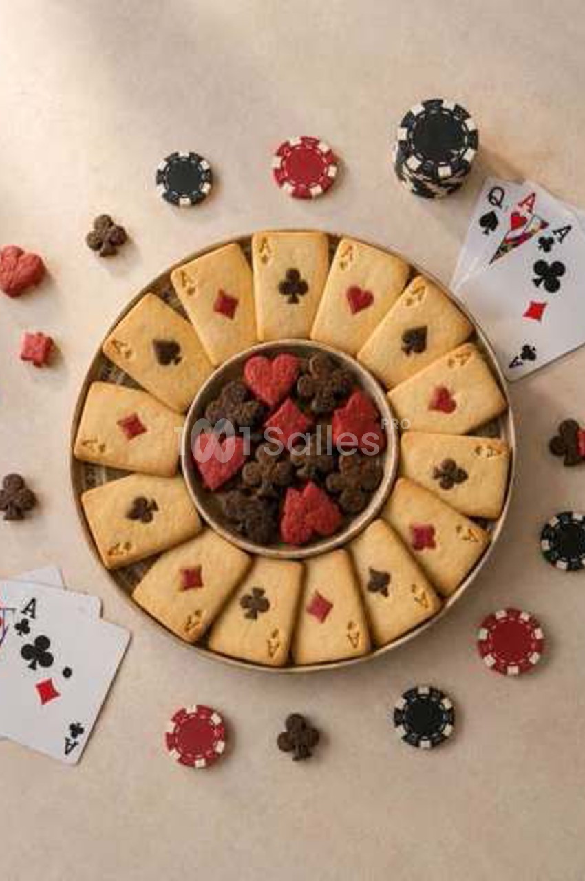 Plateau circulaire de sablés dorés en forme de cartes à jouer (as, roi, dame) ornés de symboles de pique, cœur, carreau et trèfle, entourant un bol de bonbons chocolat et fraise en forme de symboles de cartes, avec des jetons de poker disposés autour.