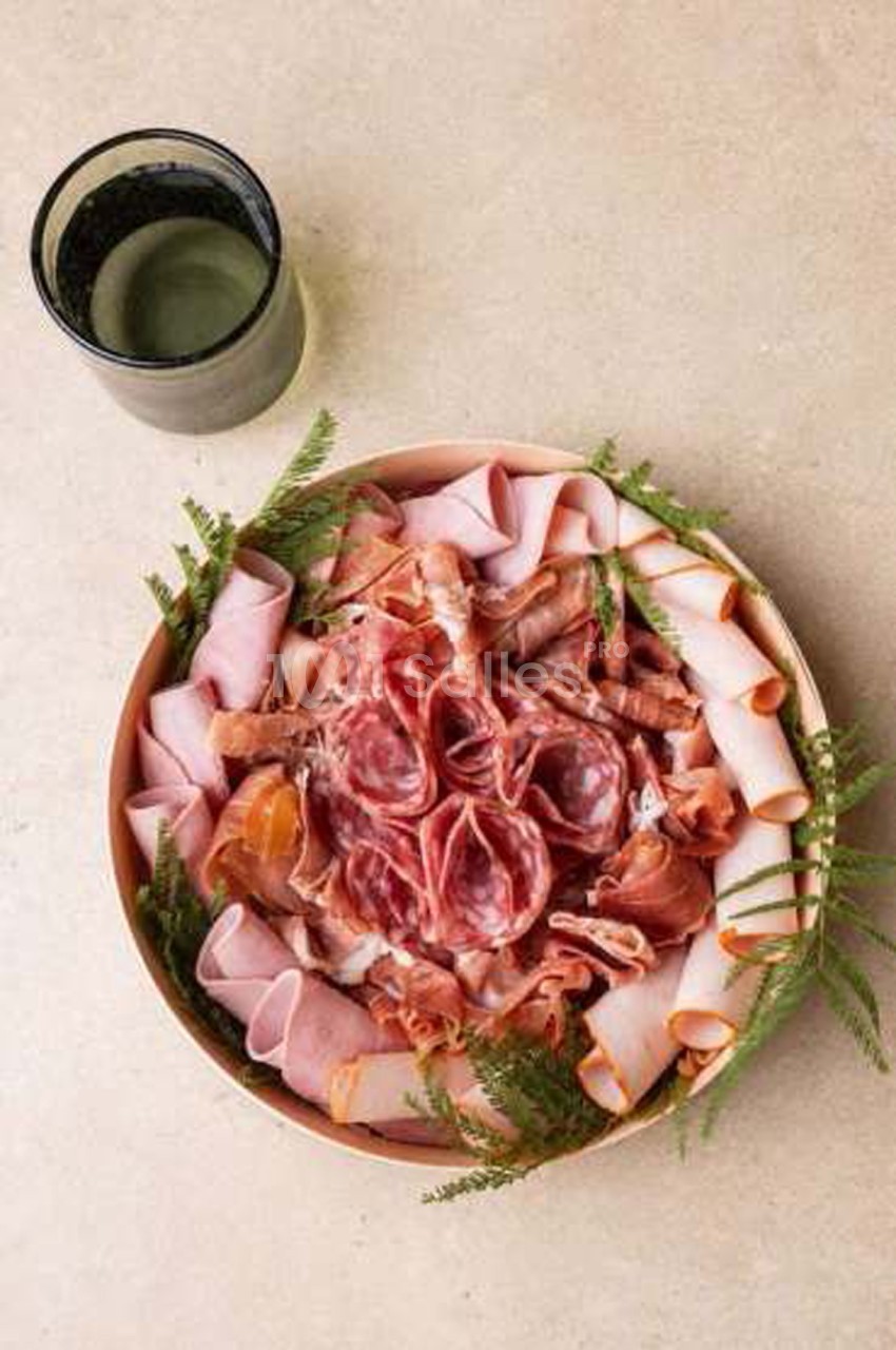 Plateau circulaire de charcuteries variées (jambon roulé, saucisson, bresaola) disposé autour d'un centre de rosettes, décoré de branches vertes, à côté d'un verre.