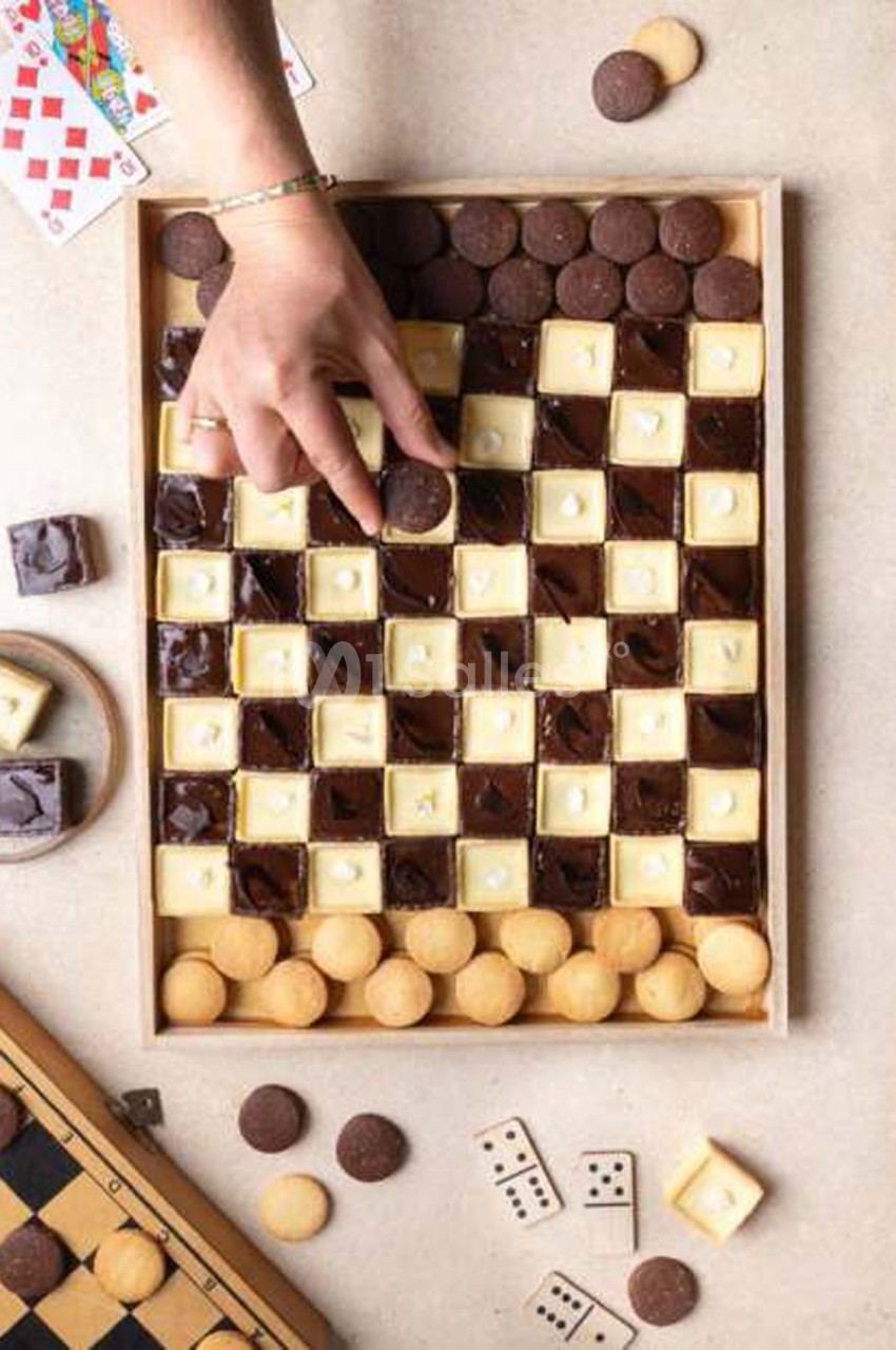 Un jeu de dames en chocolat dressé sur un plateau en bois, avec des pions en chocolat noir et blanc, et une main qui pose un pion. Let me check length: about 130 characters? Count: "Un jeu de dames en chocolat dressé sur un plateau en bois, avec des
