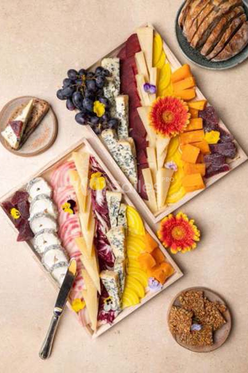 Deux planches de découpe rectangulaires présentant des fromages variés (bleus, à pâte dure, tranchés), des fruits (raisins noirs, tranches de mangue ou de pomme), des pâtes de fruits, du pain et des fleurs comestibles, sur fond beige. Let me check
