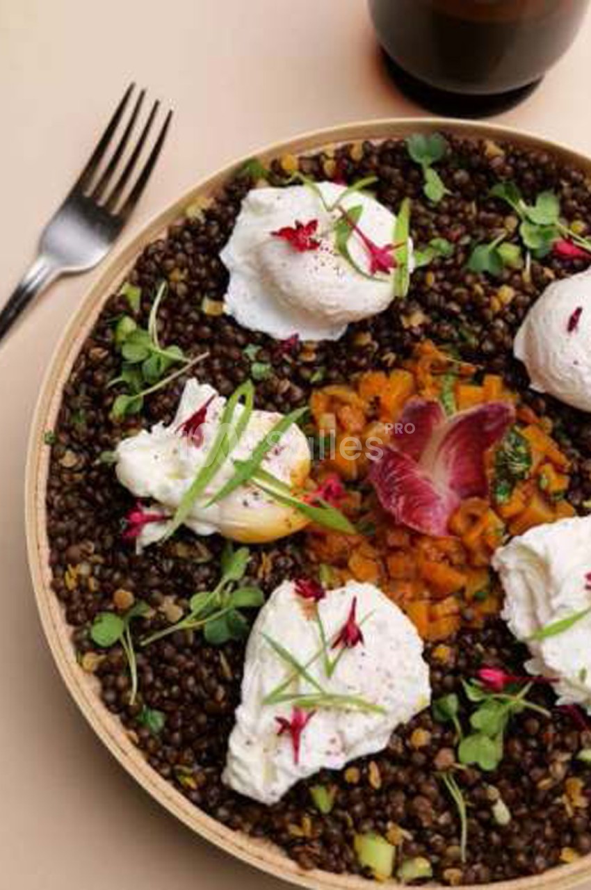 Assiette de lentilles vertes et carottes, surmontée d'œufs pochés blancs, de fleurs rouges et de pousses vertes, sur fond beige. Wait, I must check: the dish shows lentils (dark, brown/green lentils), diced carrots in orange, poached eggs (white blanc